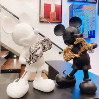 On voit deux stautes représentatn Mickey Mouse devant une scalier contemporain en verre. A droite, la première statue est blanche avec les mains argentées. La seconde, à gauche, est noire avec les mains dorées; les deux statues représentent Mickey qui forme un coeur avec ses doigts.