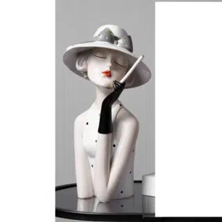 Photo d'une statue buste de femme accoudée sur une table noire portant un chapeau à larges bords avec un ruban gris et la femme un rouge à lèvres rouge, un débardeur blanc à pois gris et un gant mi-long noir qui tient un porte cigarette style année 20