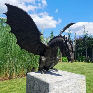 Photo d'un dragon gris foncé ailes déployées tête baissée assis sur un cube de pierre dans un jardin