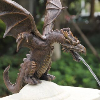 Photo d'un dragon ailé gothique cuivré posé sur un monticule en pierre blanche et crachant de l'eau dans un jardin flouté à l'arrière plan