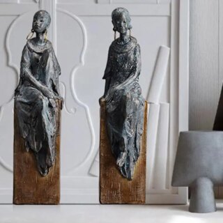 Photo de deux statues africaines de femmes l'une tête baissée , l'autre tête relevée assises sur un socle marron et portant une robe longue fluide, un large collier et des grandes boucles d'oreilles le tout dans un décor épuré contemporain blanc