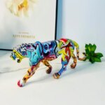 Sur un meuble blanc moderne, on voit une statue de panthère moderne avec un motif graffiti coloré. Elle est près d'un cadre avec une fleur.