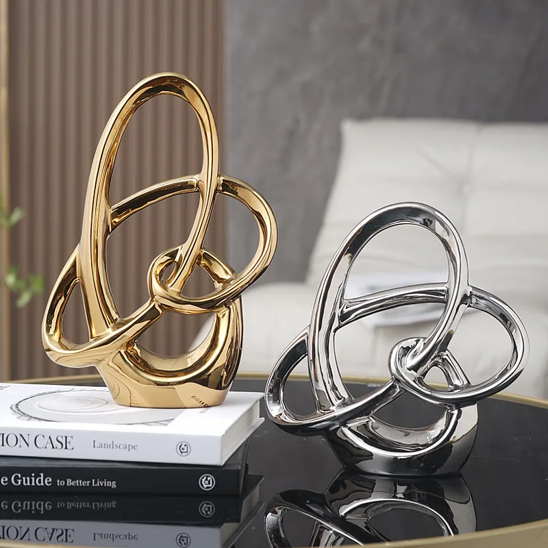 2698-uamyvi.jpg photo de deux Statue géométrique en céramique et émaille formant des anneaux entre méllés, une dorée et une argentée, posée sur un bureau en verre noir avec une chaise blanche derrière