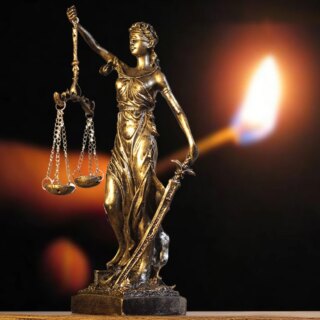 Photo d'une statue dorée grecque de Thémis la déesse de la justice avec la balance dans la main droite et le glaive dans la main gauche sur un petit promontoire en arrière fond flou une allumette qui brûle