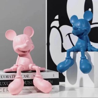 On voit deux statues de Mickey, une rose et une bleue, assises sur un meuble et une pile de livres.