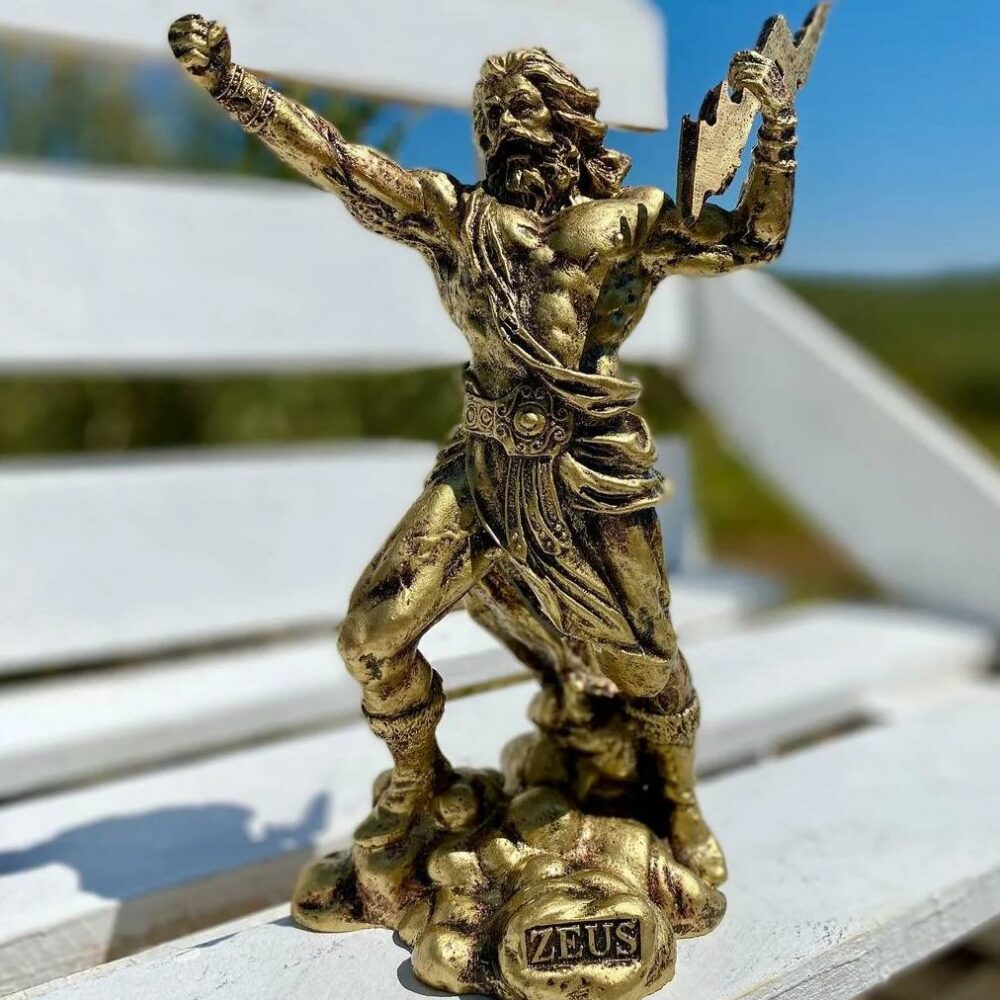 3947-6b2f7d.jpeg Sur un banc blanc, en extérieur, on voit une statue de Zeus, dorée, qui lève le poing au ciel et qui tient un éclair dans l'autre main. Il s'apprête à partir en guerre.