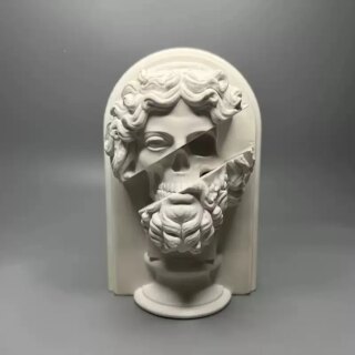 On voit, sur fond gris une statue en plâtre de Zeus. Son visage est creusé au milieu et laisse apparaître son squelette.