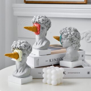 3 statues street art buste de David avec un cornet de glace sur le visage, posée sur des libres sur une table blanche.