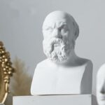 On voit une statue, le buste d'un homme : Socrate. Elle est posée sur un meuble blanc devant un décor gris et minimaliste.