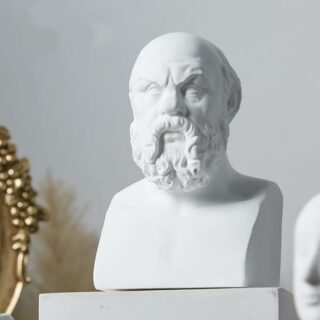 On voit une statue, le buste d'un homme : Socrate. Elle est posée sur un meuble blanc devant un décor gris et minimaliste.