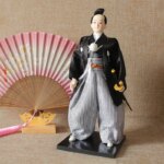 Devant un éventail rose japonais, on voit la statue d'un samouraï japonais en costume traditionnel. Il porte un sabre à la taille.