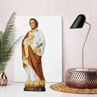 Photo d'une statue de Saint Joseph aux cheveux et barbe roux souriant vêtu d'une robe blanche aux pourtours dorés et d'une étoffe marron par dessus pieds nus et tenant en mains une grande tige de fleurs et ungenre d'équerre dorée le tout sur un double socle couleur pierre et le second noir avec son nom gravé la statue est sur un meuble blanc entourée d'un luminaire et d'une plante