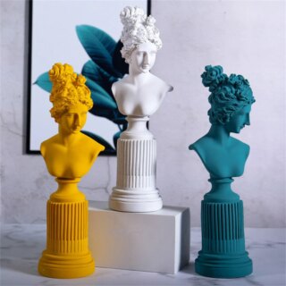 Photo de trois statues de bustes de femmes à chignons seins nus sur colonne de trois couleurs une jaune, une bleue, et une blanche sur un cube blanc