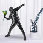 Statue street art Banksy lanceur de fleurs noire posée sur un meuble blanc à côté de fleurs bleues