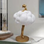 Photographie d'une statue représentant une jeune femme dorée dans un nuage blanc en train de danser la tête relevée le tout sur un socle rond lui-même posé sur un meuble style marbre