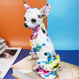 On voit une statue de chien type Chihuahua blanche avec des éclats de couleurs vives. Elle est en résine et posée sur un livre devant un mur orange et bleu.