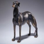 On voit une statue de lévrier en bronze sur fond gris dégradé.