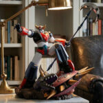 Cette statue Goldorak triomphant d'un golgoth représente le célèbre robot géant brandissant sa hache devant un fauteuil en cuir, avec une bibliothèque en arrière-plan. La figurine colorée écrase un ennemi mécanique sous son pied dans une pose de victoire héroïque.